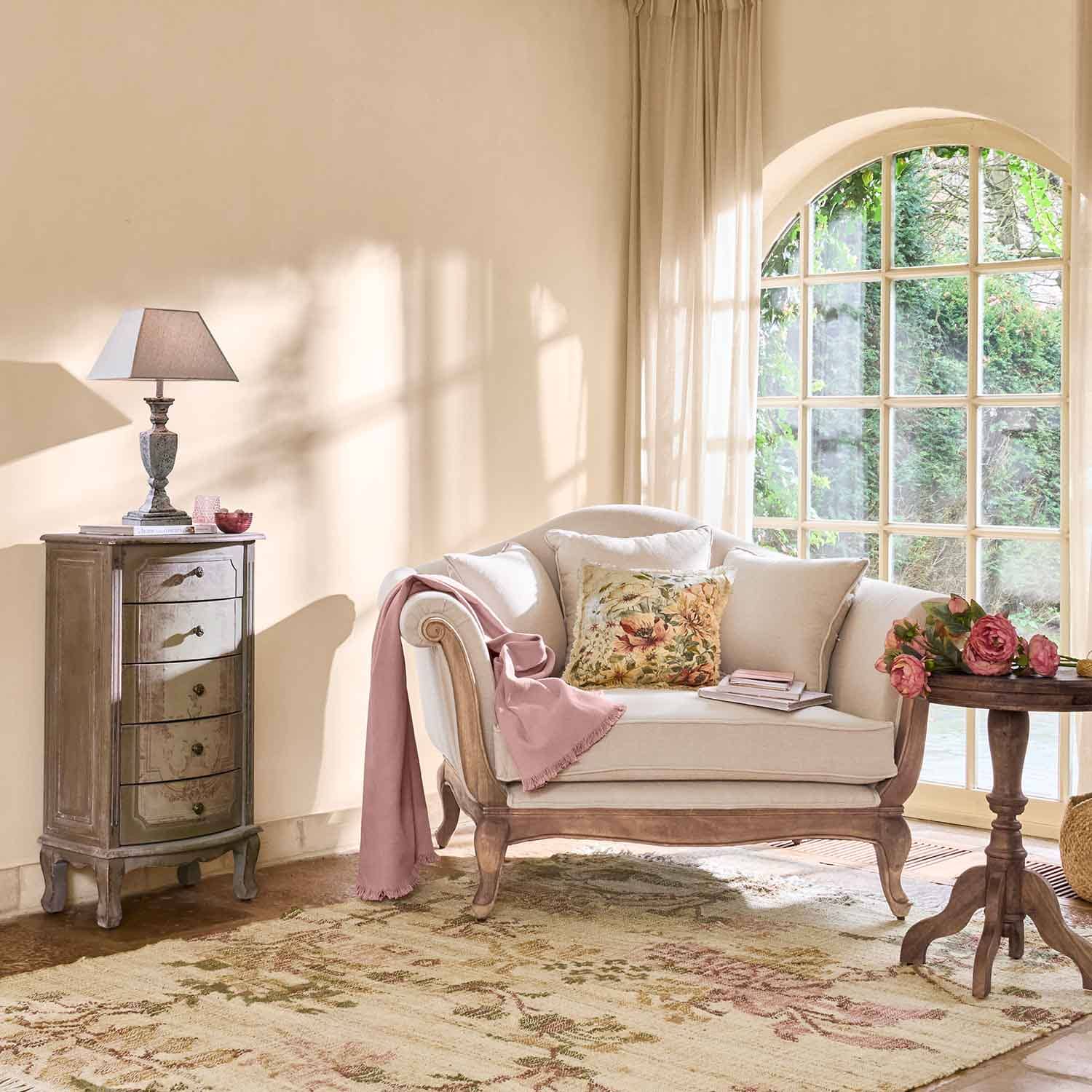 Cremefarbige Sofa mit Holzrahmen, Blumenmuster-Kissen und rosa Decke in hellem Wohnzimmer mit großem Fenster