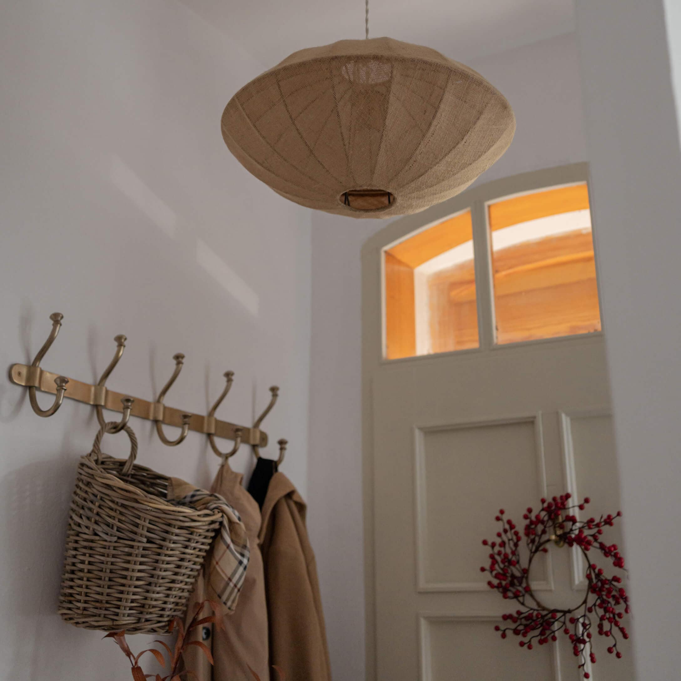 Beige Stoff-Hängeleuchte in schirmartigem Design über Garderobe mit Korb und roten Beeren an weisser Tür