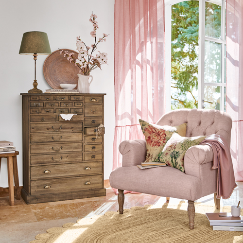 Gemütlicher rosa Sessel mit Blumen-Kissen neben einer Holzkommode im hellen Wohnzimmer mit rosa Vorhängen