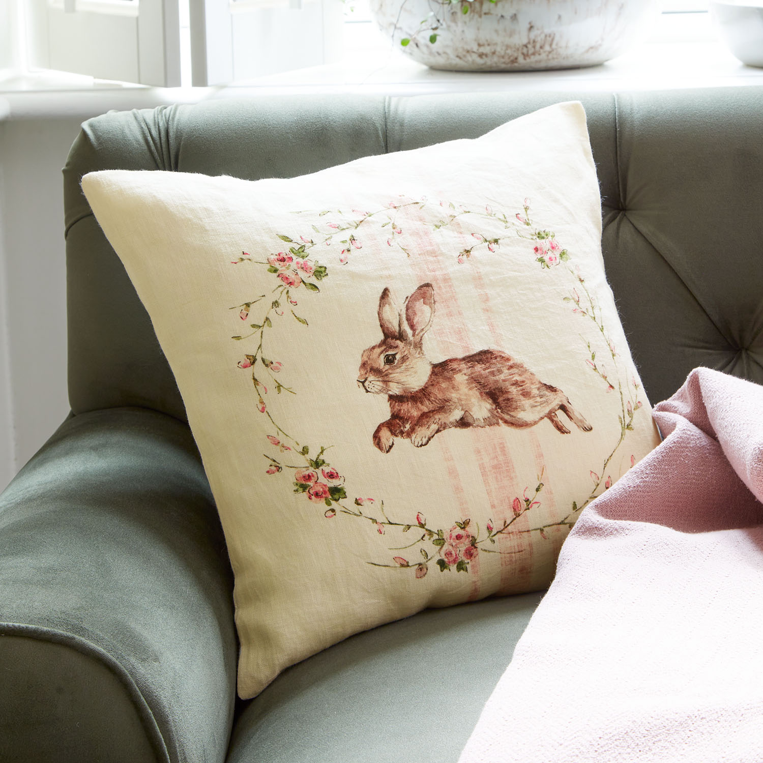 Beiges Leinenkissen mit Hasenmotiv und Blumenranken auf grünem Sofa mit rosa Decke