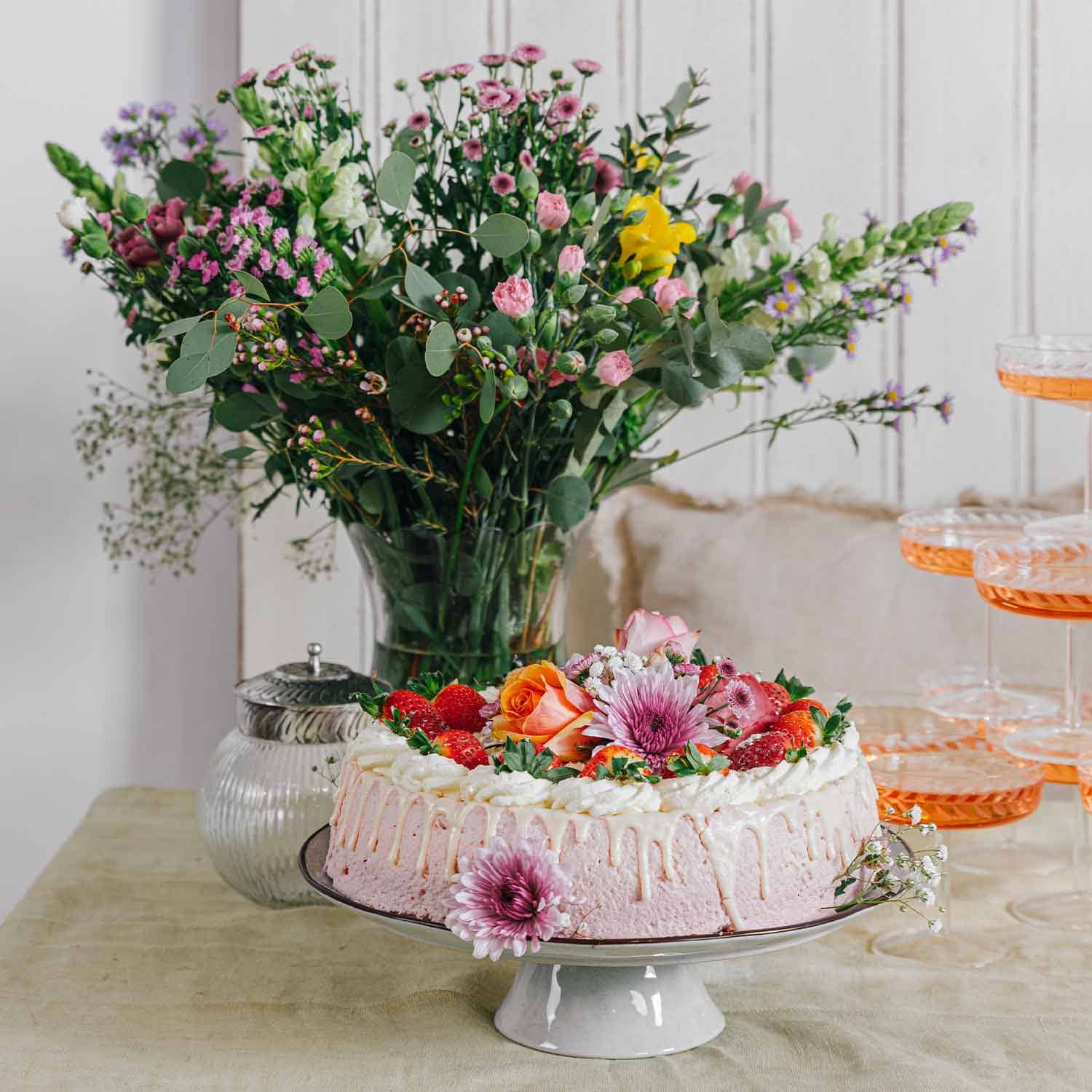 Rosafarbene Sahnetorte mit frischen Erdbeeren und Blumen auf weissem Tortenständer vor Blumenstrauss