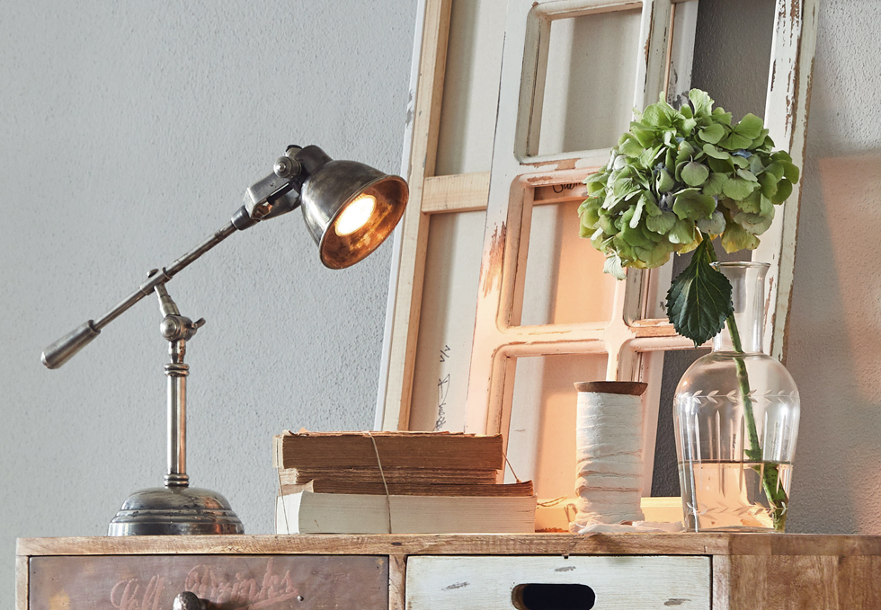 Industrielle Metall-Schreibtischlampe auf Holzkommode mit Büchern und Vase mit grünem Blumenstrauss