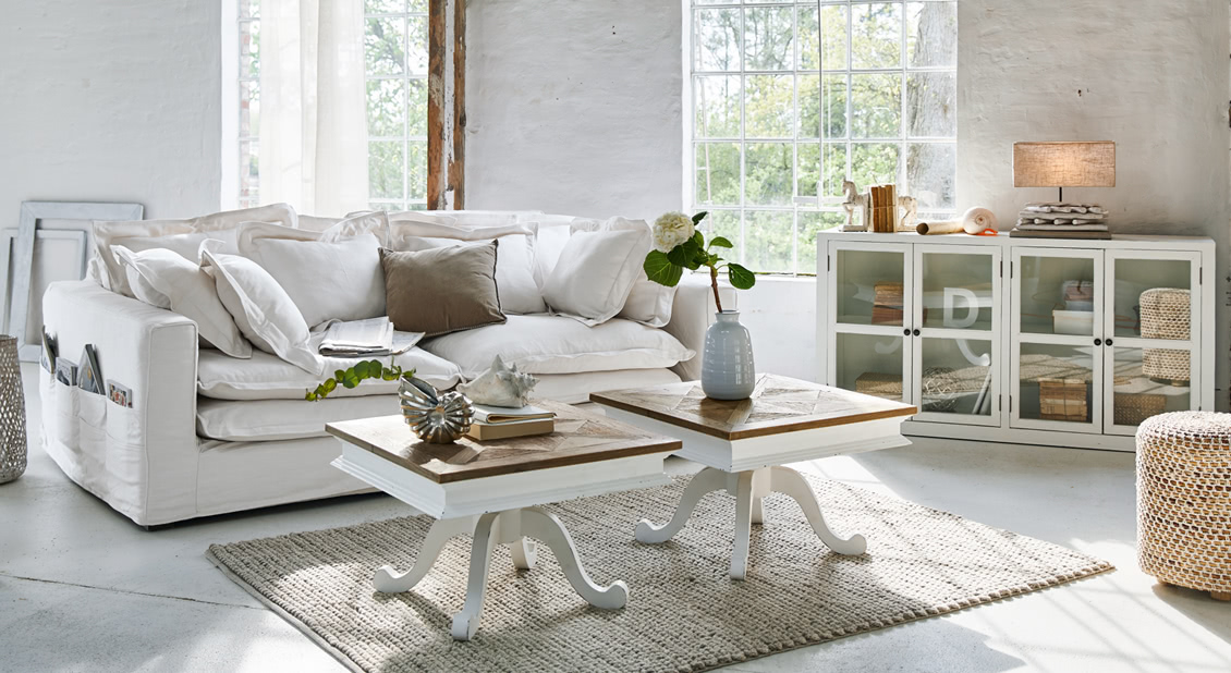 Helles Wohnzimmer mit weissem Sofa, zwei Holzcouchtischen und hellem Teppich im Landhausstil