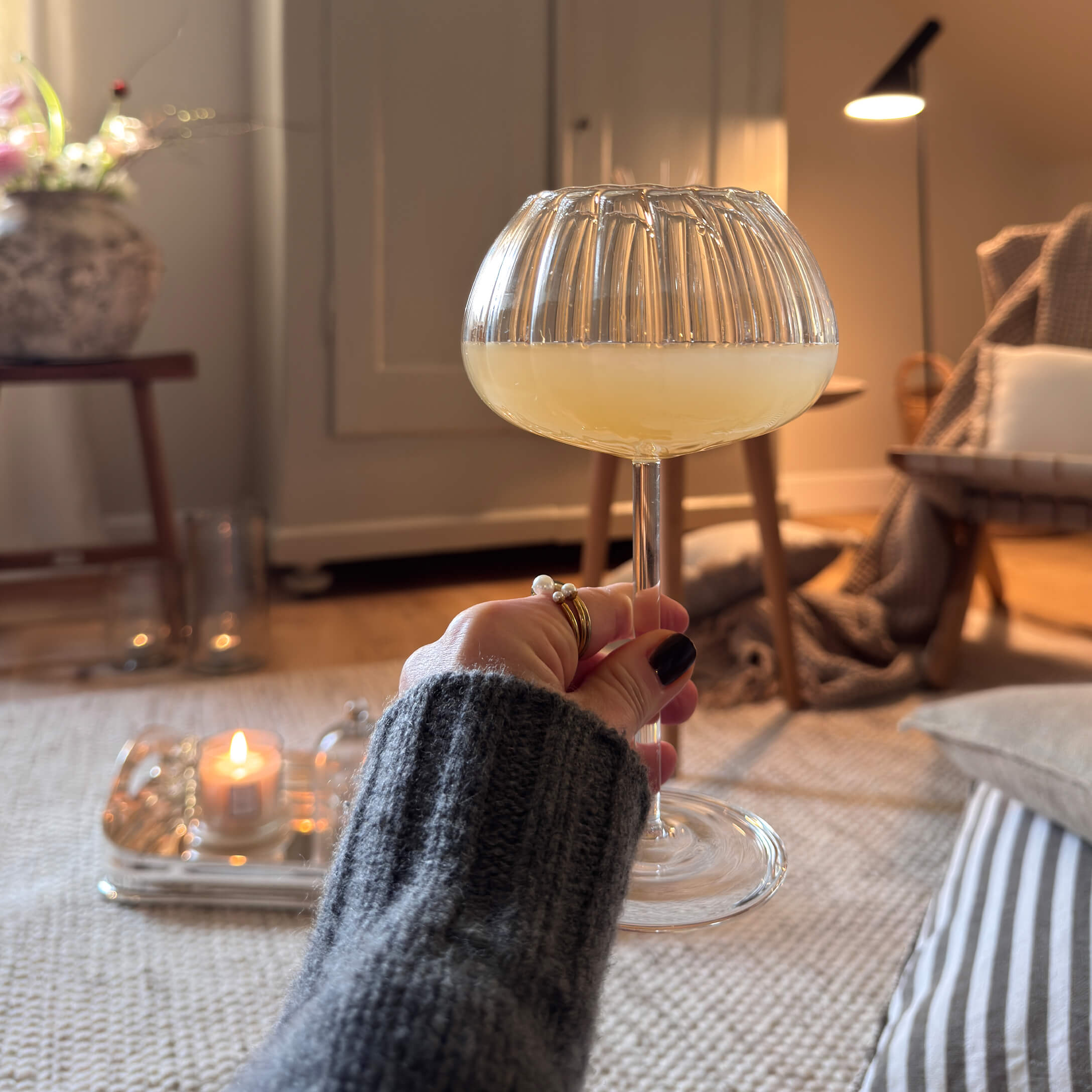 Hand hält ein Glas mit gelbem Getränk in einem gemütlichen Raum mit sanfter Beleuchtung und Dekorationen.