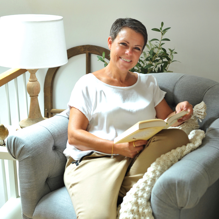 Frau in weissem Oberteil und beiger Hose liest Buch auf grauem Sofa in hellem Zimmer