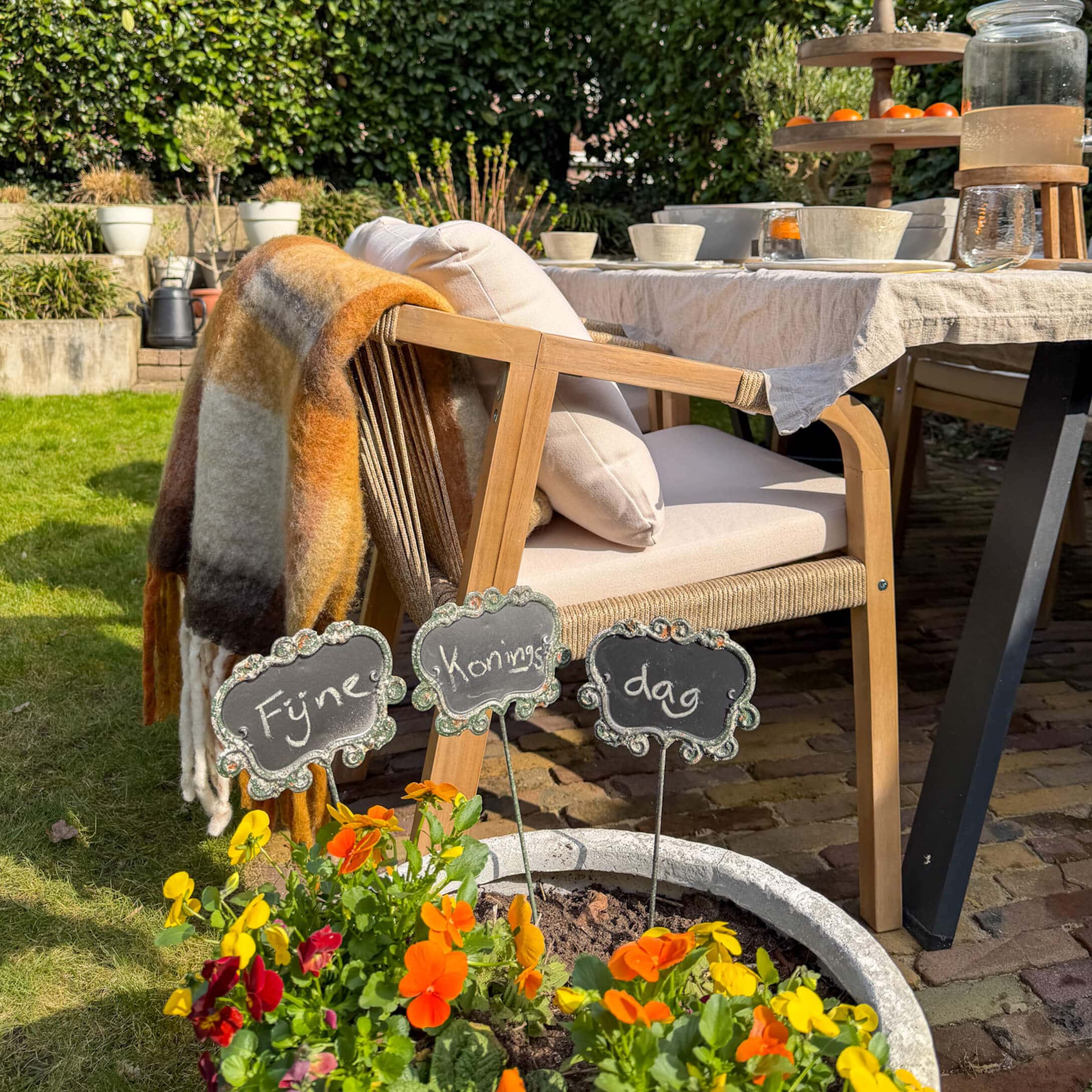 Holz-Gartenstuhl mit hellen Kissen und buntem Plaid neben gedecktem Tisch und bunten Blumen im Topf