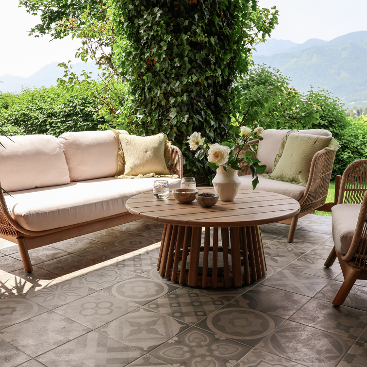 Gartenmöbel aus Rattan mit beigen Kissen und rundem Holztisch auf Terrasse mit gemusterten Platten