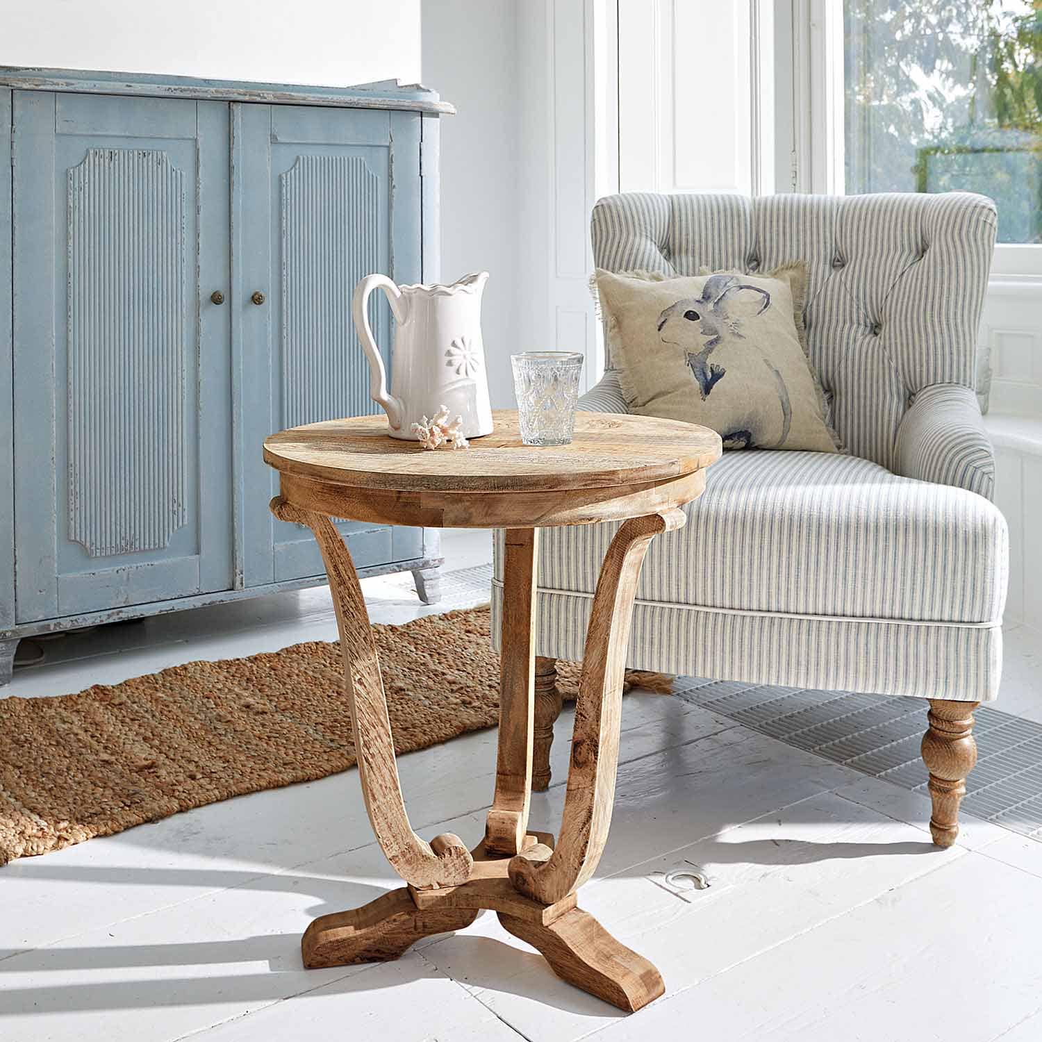 Runder Beistelltisch aus Mangoholz in Naturfarbe im hellen Wohnzimmer mit gestreiftem Sessel und blauem Schrank