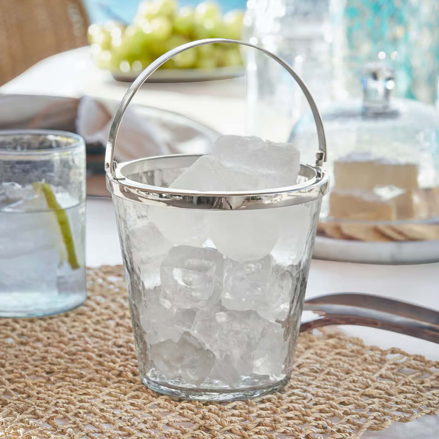 Transparentes Glas-Eiswürfeleimer mit Metallgriff auf Tisch mit geflochtenem Untersetzer