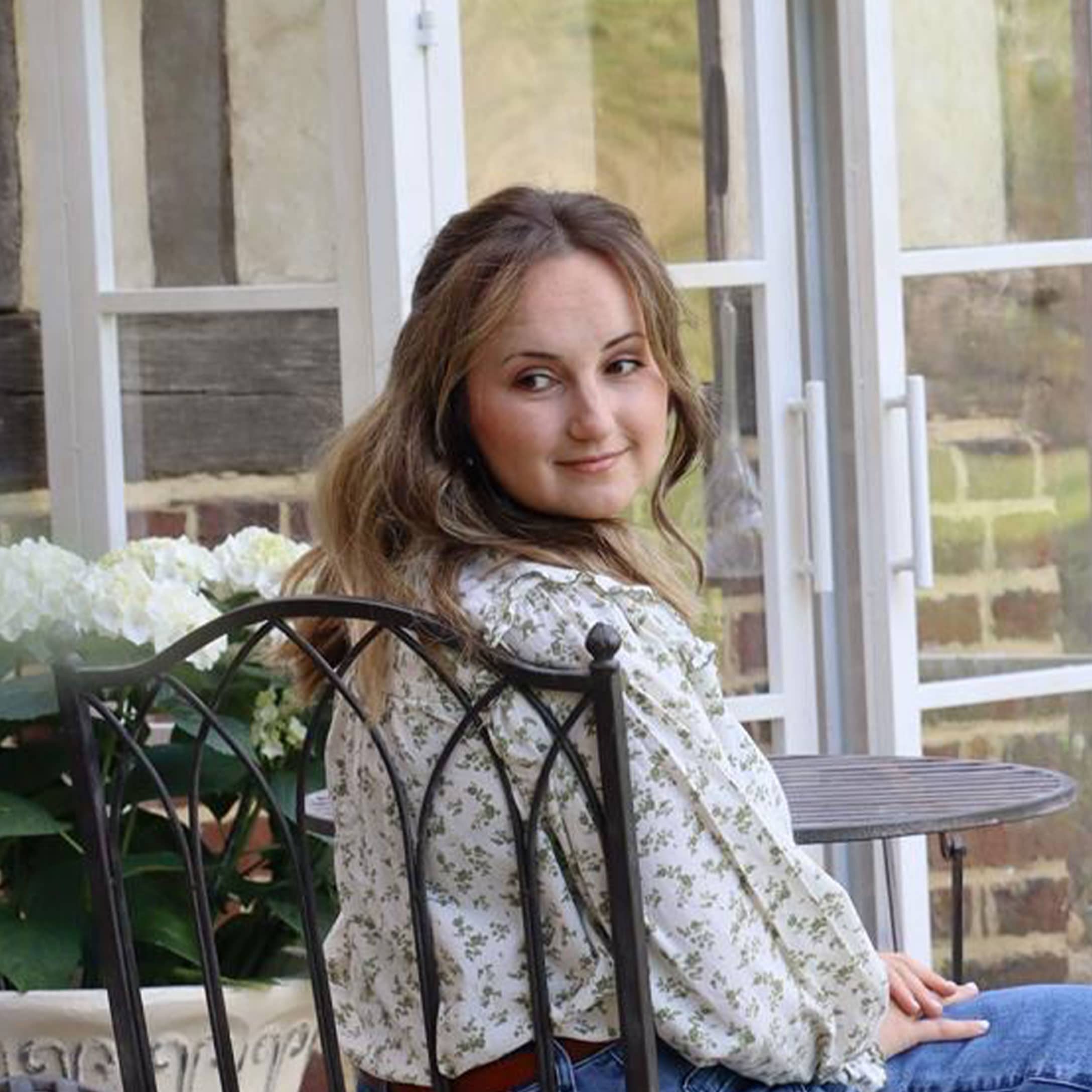 Frau in geblümter Bluse sitzt auf Metallstuhl in hellem Garten mit weissen Blumen