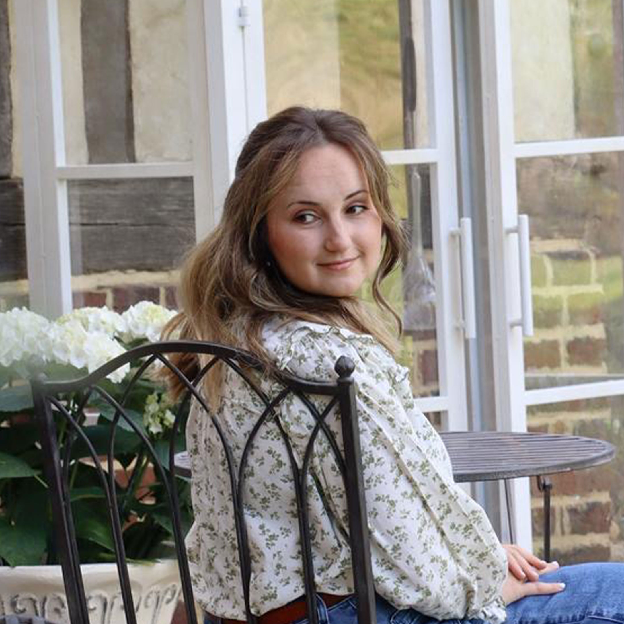Frau in geblümter Bluse sitzt auf Metallstuhl in hellem Garten mit weissen Blumen