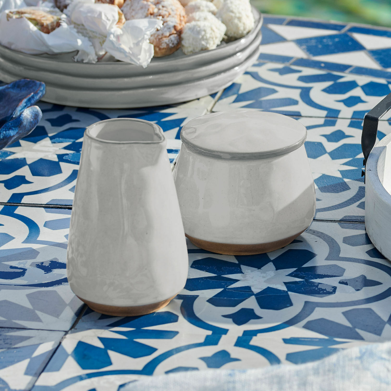 Zuckerdose und Milchkännchen Biarré Weisse Keramikkanne und Zuckerdose mit braunem Naturboden auf blau-weiss gefliestem Tisch