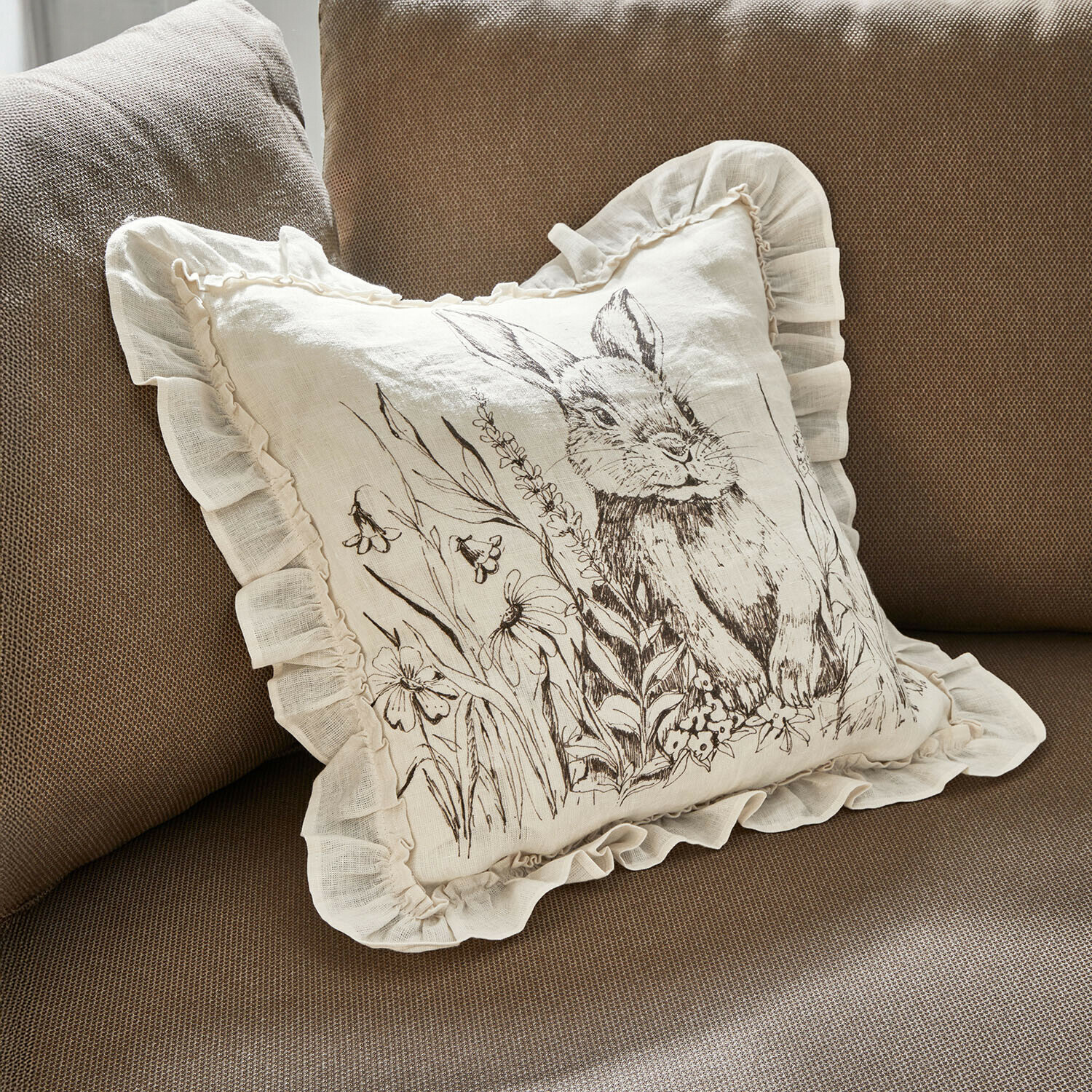 Kissenhülle Brionelle Beiges Leinenkissen mit Rüsche und Hasenmotiv mit Blumen auf braunem Sofa