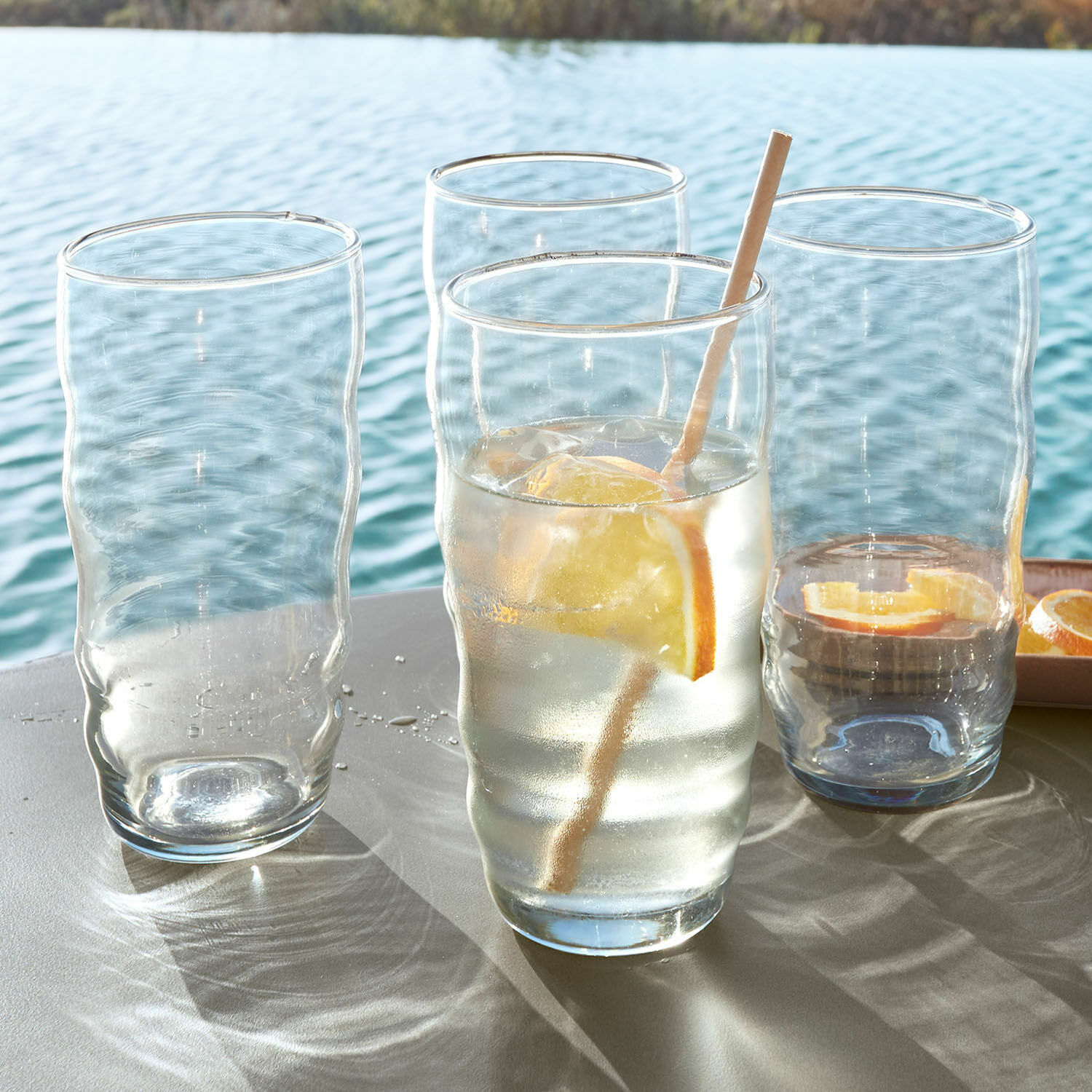 Glas 4er Set Tournavin Vier durchsichtige, wellenförmige Gläser auf einem Tisch am Wasser, ein Glas mit Zitrone und Strohhalm.