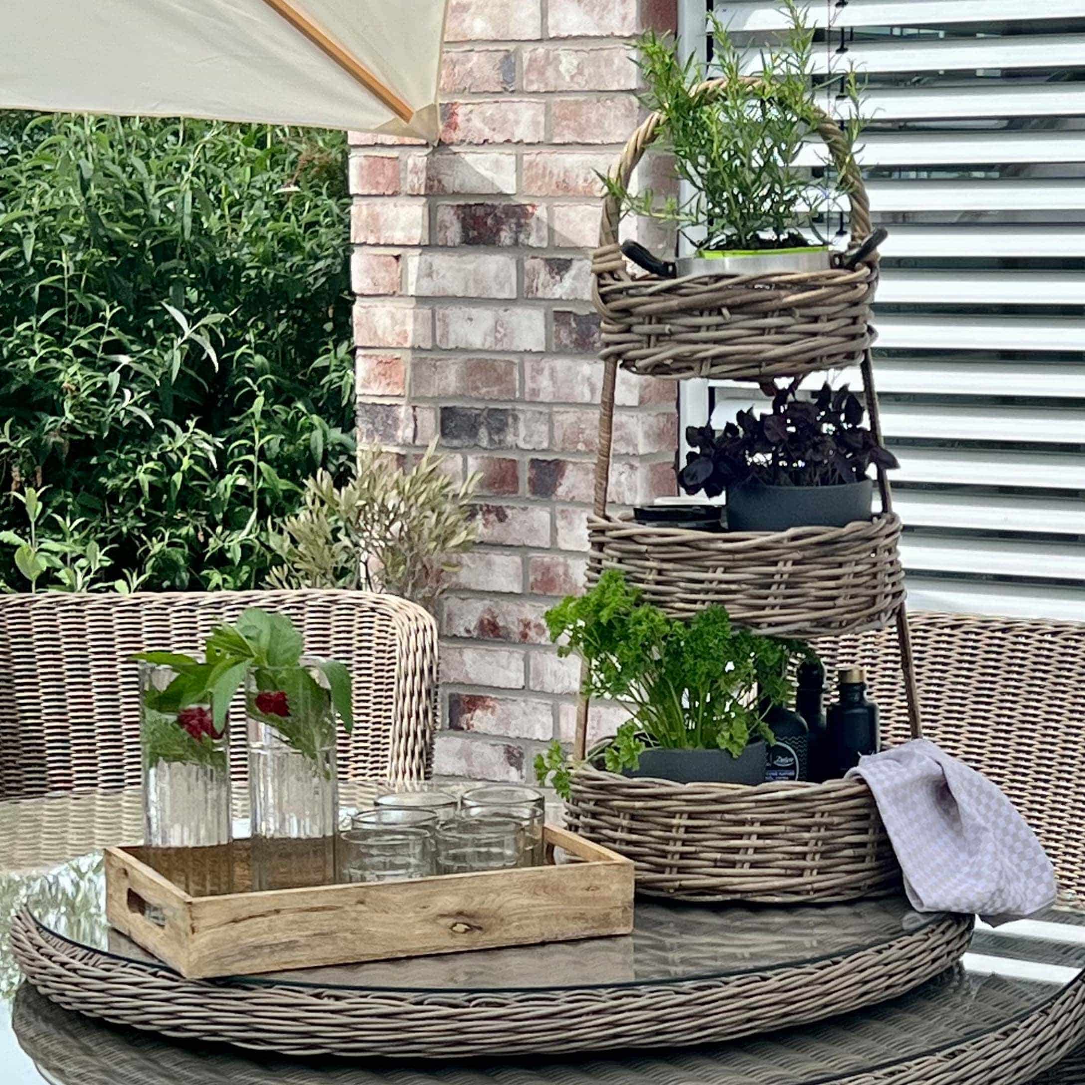 Dreistöckiger Rattanständer mit grünen Pflanzen auf Glastisch auf Terrasse mit Rattanmöbeln