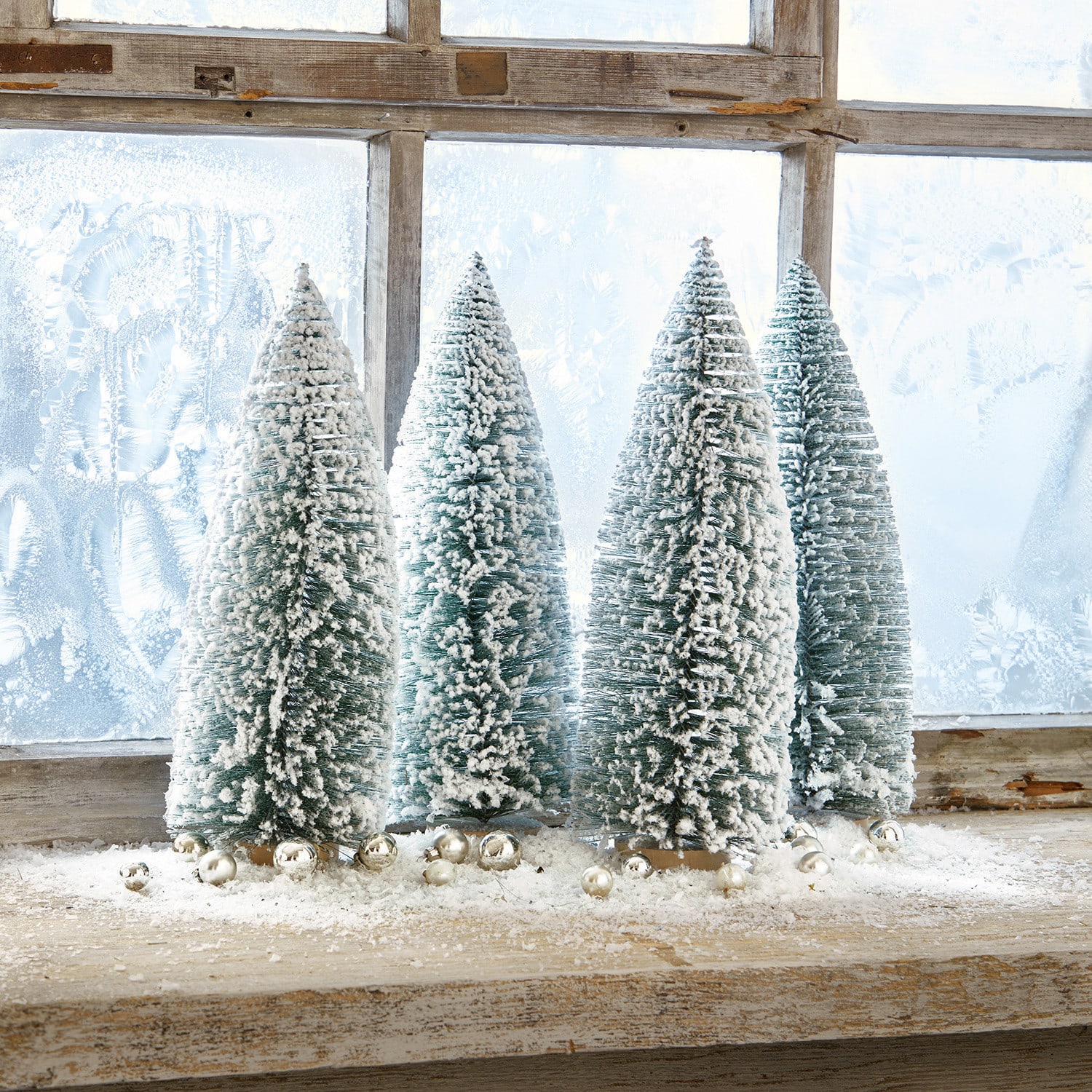 Deko-Baum 4er Set Landscape Vier grüne Kunststoff-Tannenbäume in Kegelform mit weissem Schnee auf einem Holzfensterbrett mit silbernen Kugeln