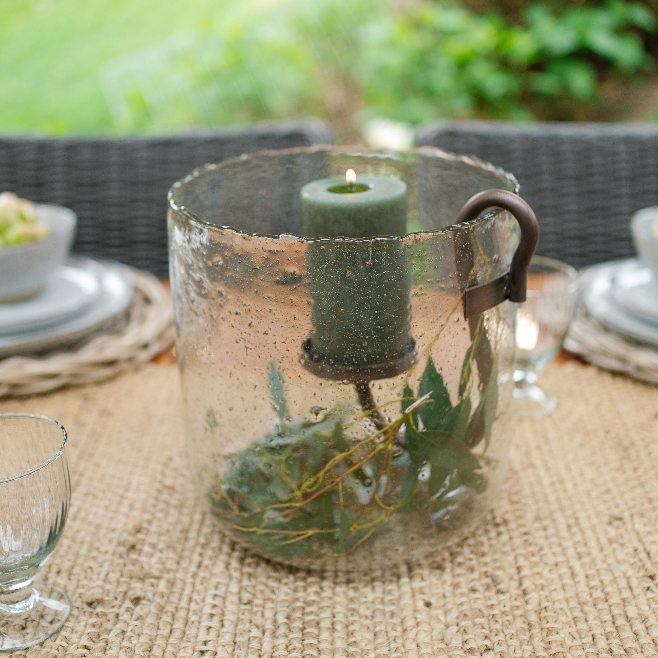 Glaswindlicht mit grüner Kerze und Metallgriff auf rustikalem Gartentisch