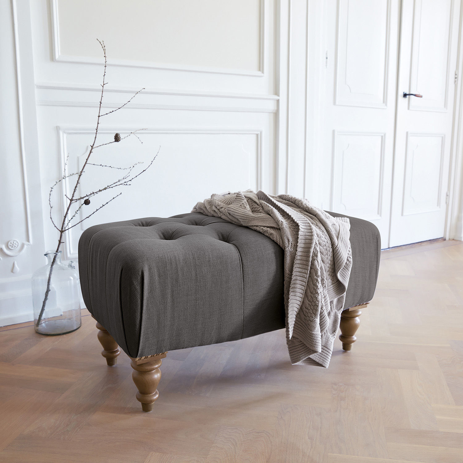 Hocker Durance Grau gepolsterter Hocker mit Holzbeinen und beiger Decke in hellem Raum
