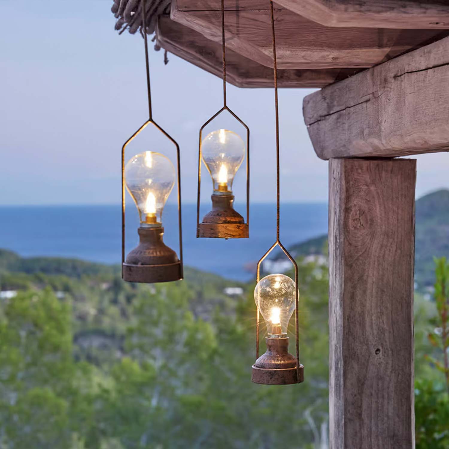 Drei hängende Metalllampen mit Glühbirnen vor grünem Wald und Meer, Aussenbeleuchtung