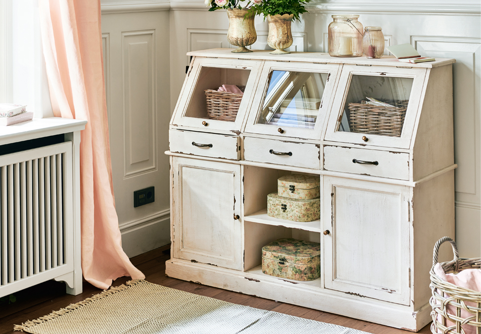 Weisser Holzschrank im Shabby-Chic-Stil mit Glastüren und Dekorationen in einem hellen Raum.