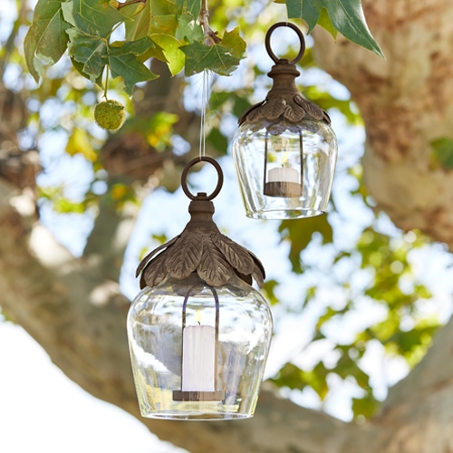 Glaslaternen mit Metallverzierung und weissen Kerzen hängen an einem Baum als Gartendekoration.