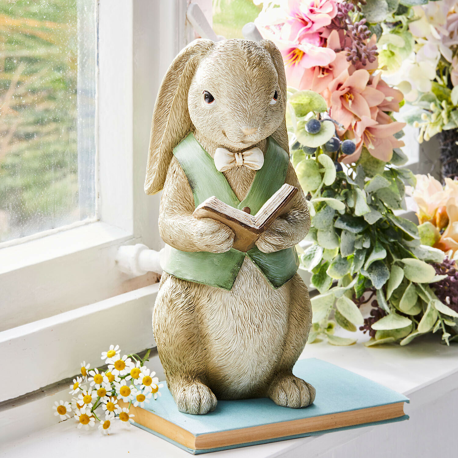 Hasenfigur aus Kunstharz mit grünem Gilet und Buch auf Fensterbank mit Blumen