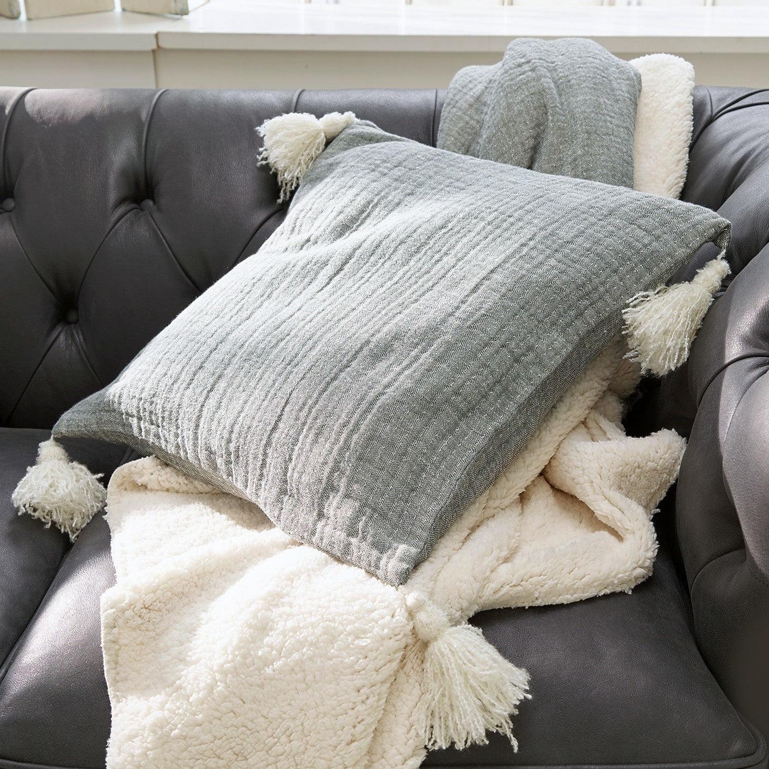 Kissen Malliévre Graues Kissen mit Quasten und cremefarbene, weiche Decke auf dunklem Ledersofa im Wohnzimmer