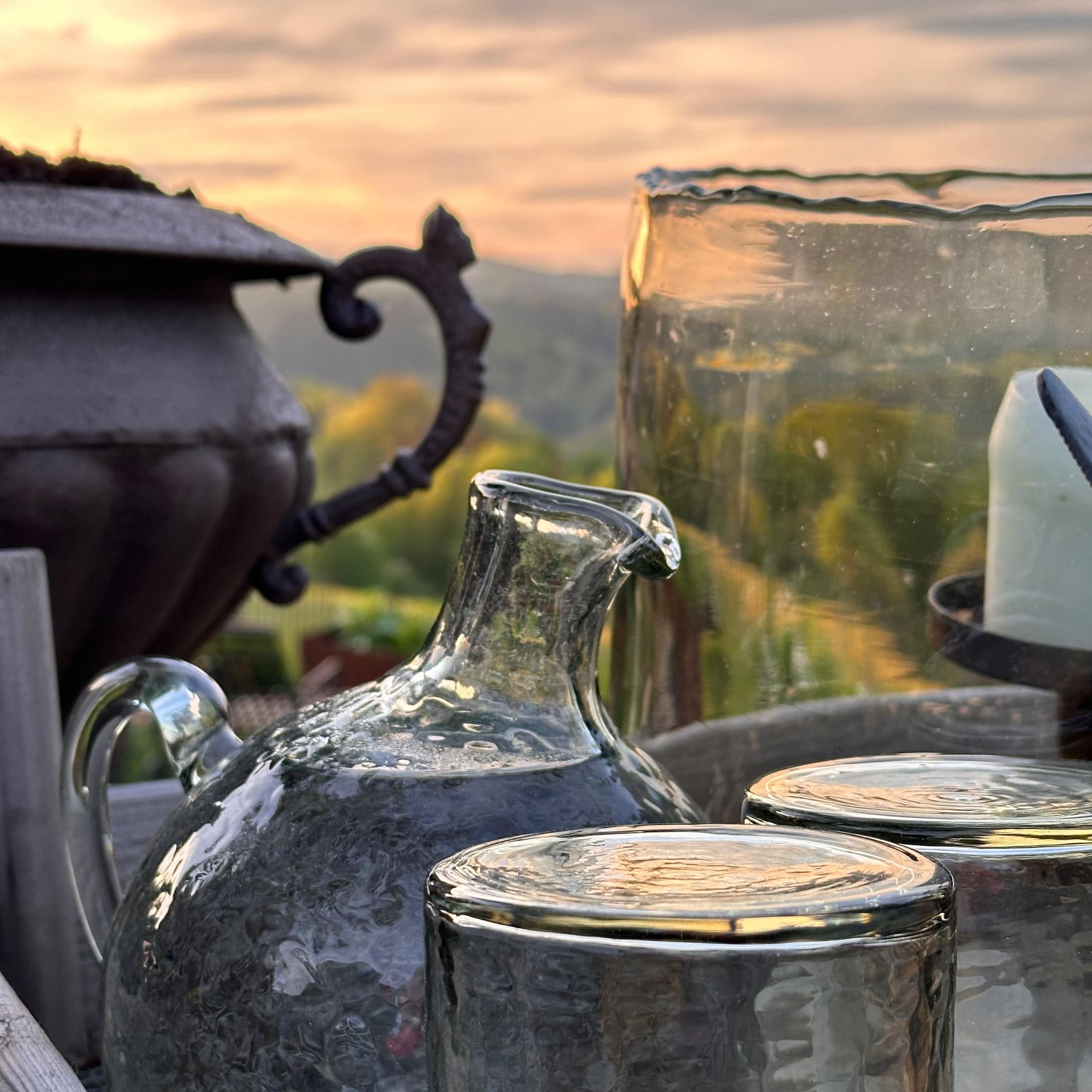 Glaskrug und Gläser mit Struktur vor sommerlichem Garten bei Sonnenuntergang