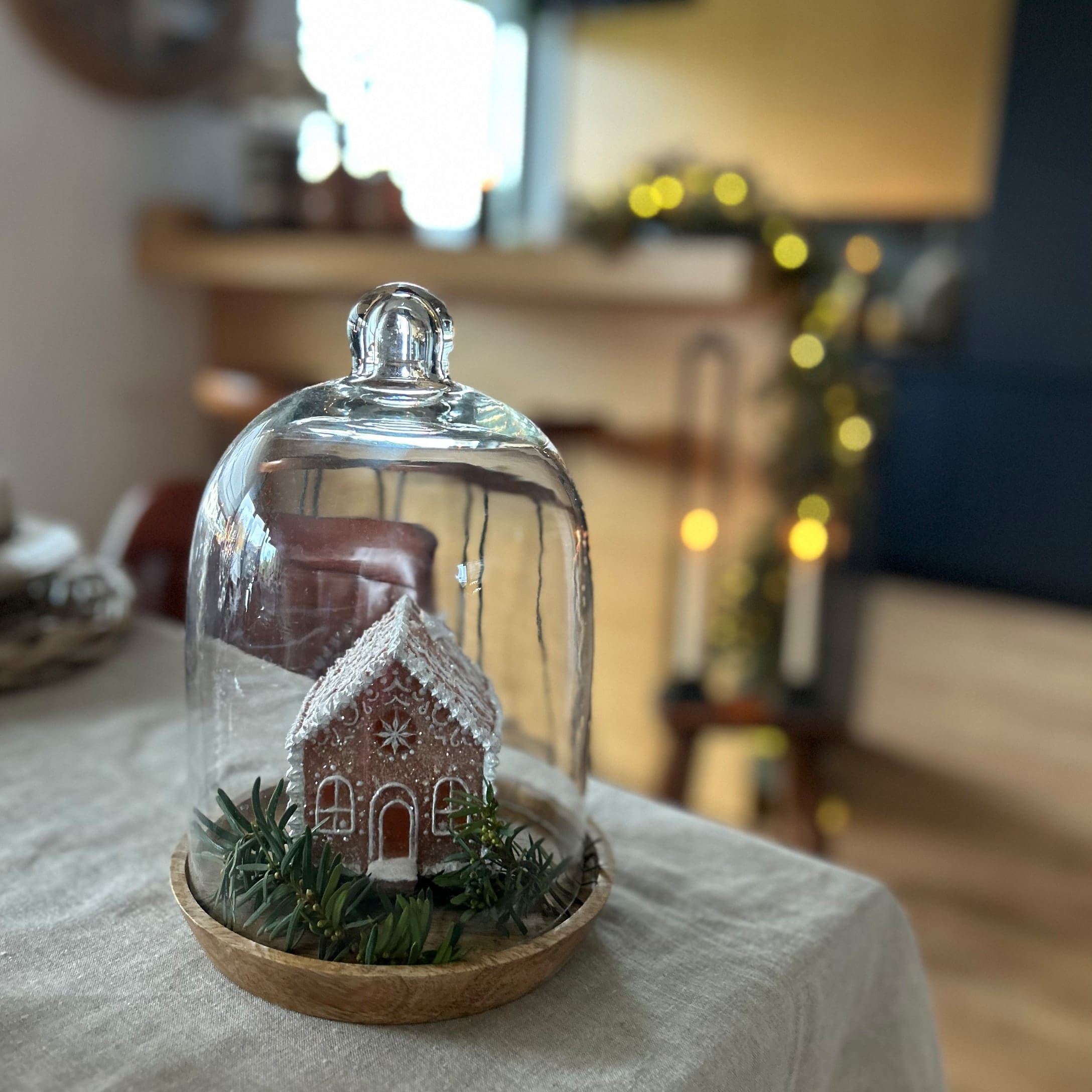 Dekoratives Lebkuchenhaus unter Glaskuppel auf Holzplatte mit grünen Zweigen als Dekoration