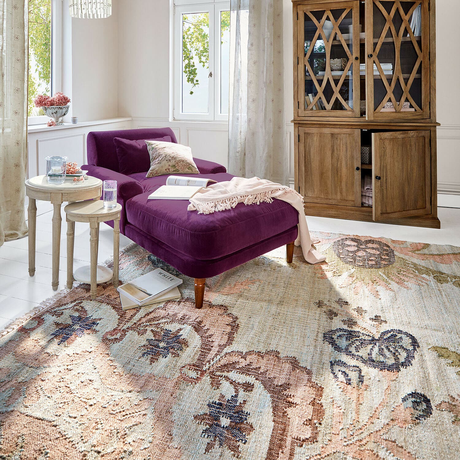 Teppich Amélorie Violette Samt-Chaiselongue mit Holzbeinen auf gemustertem Teppich in hellem Wohnzimmer mit Holzvitrine