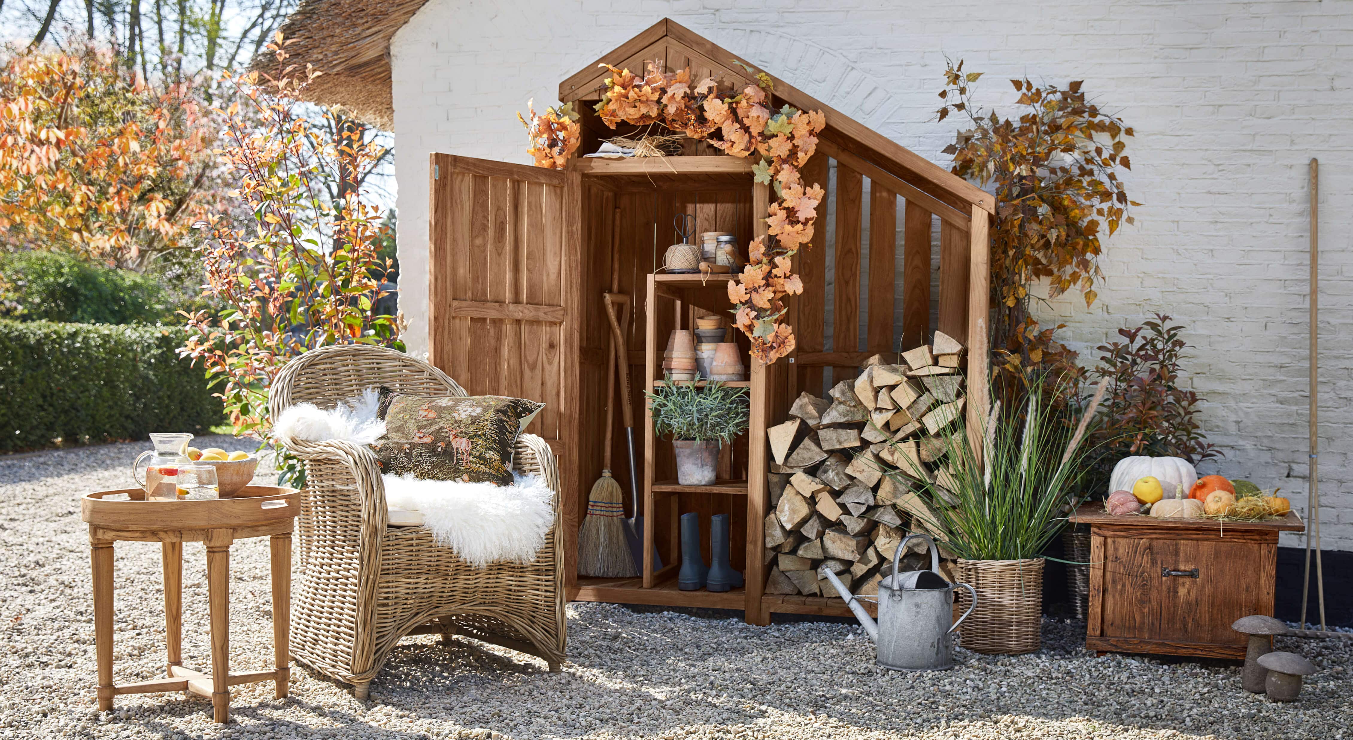 Holz-Gartenschuppen mit Herbstdekoration, Rattansessel und Beistelltisch mit Karaffe auf Kiesboden