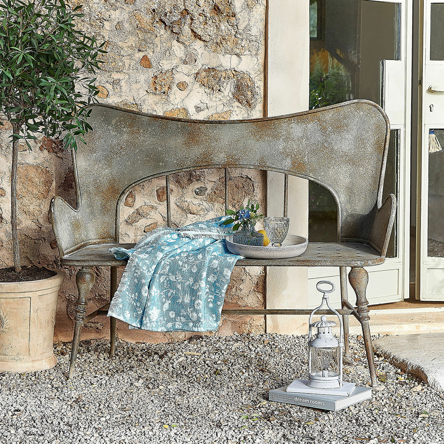 Bank Horivel Vintage-Metallbank mit grauer Patina und blauem Stoff auf Kies vor einer Steinmauer im Garten