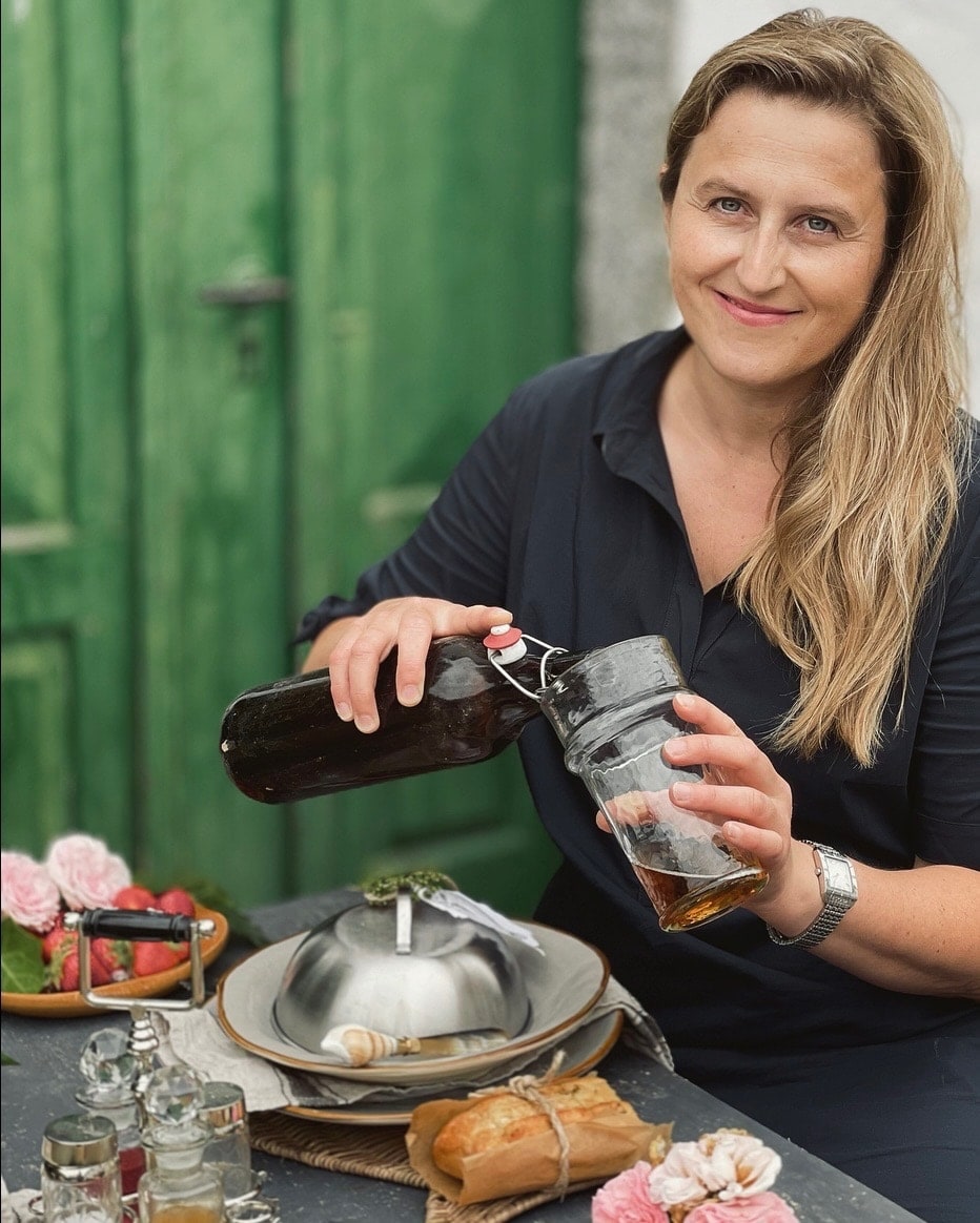 Eine Frau gießt ein Getränk aus einer Glasflasche in ein Glas am Tisch mit Essen und Blumen.