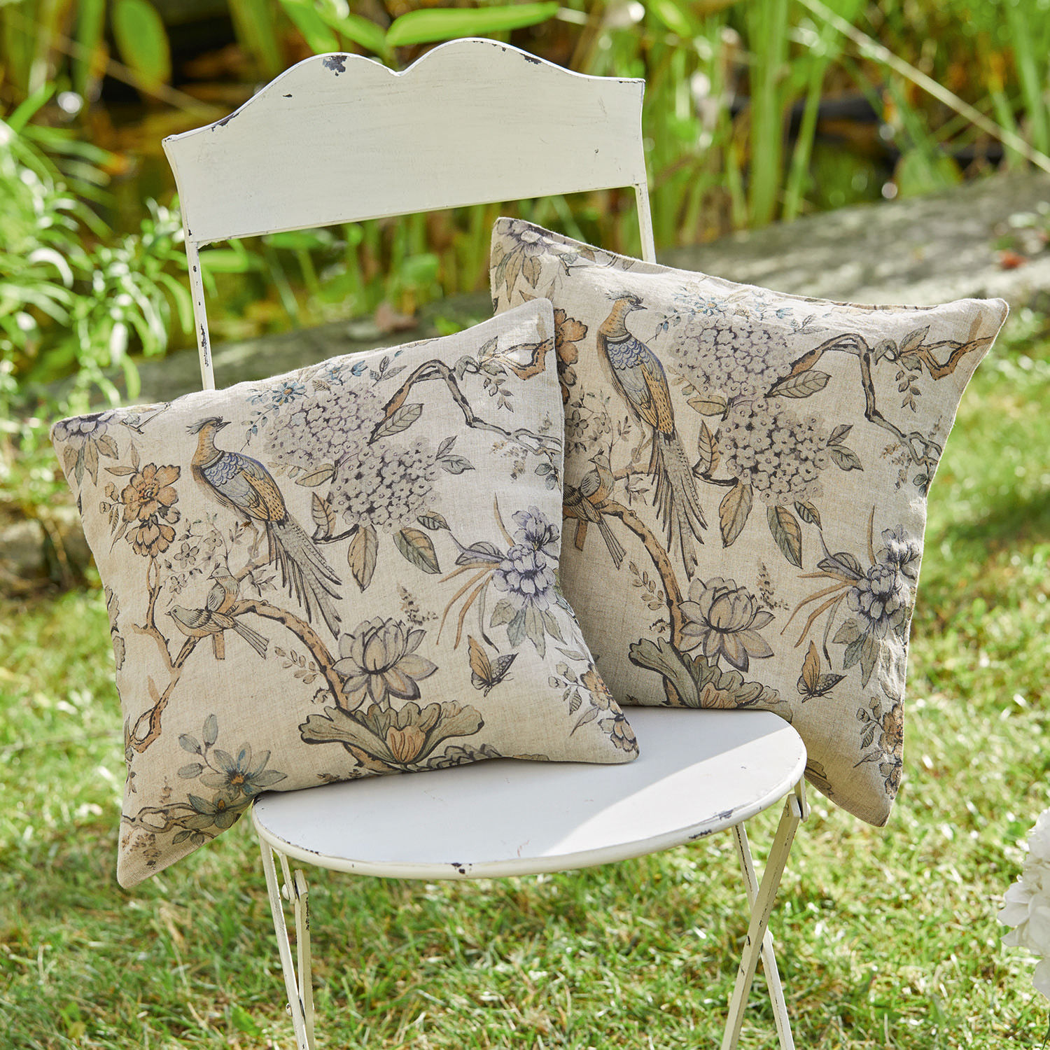 Zwei dekorative Baumwollkissen in Beige mit Vogel- und Blumenmuster auf weissem Stuhl im Garten