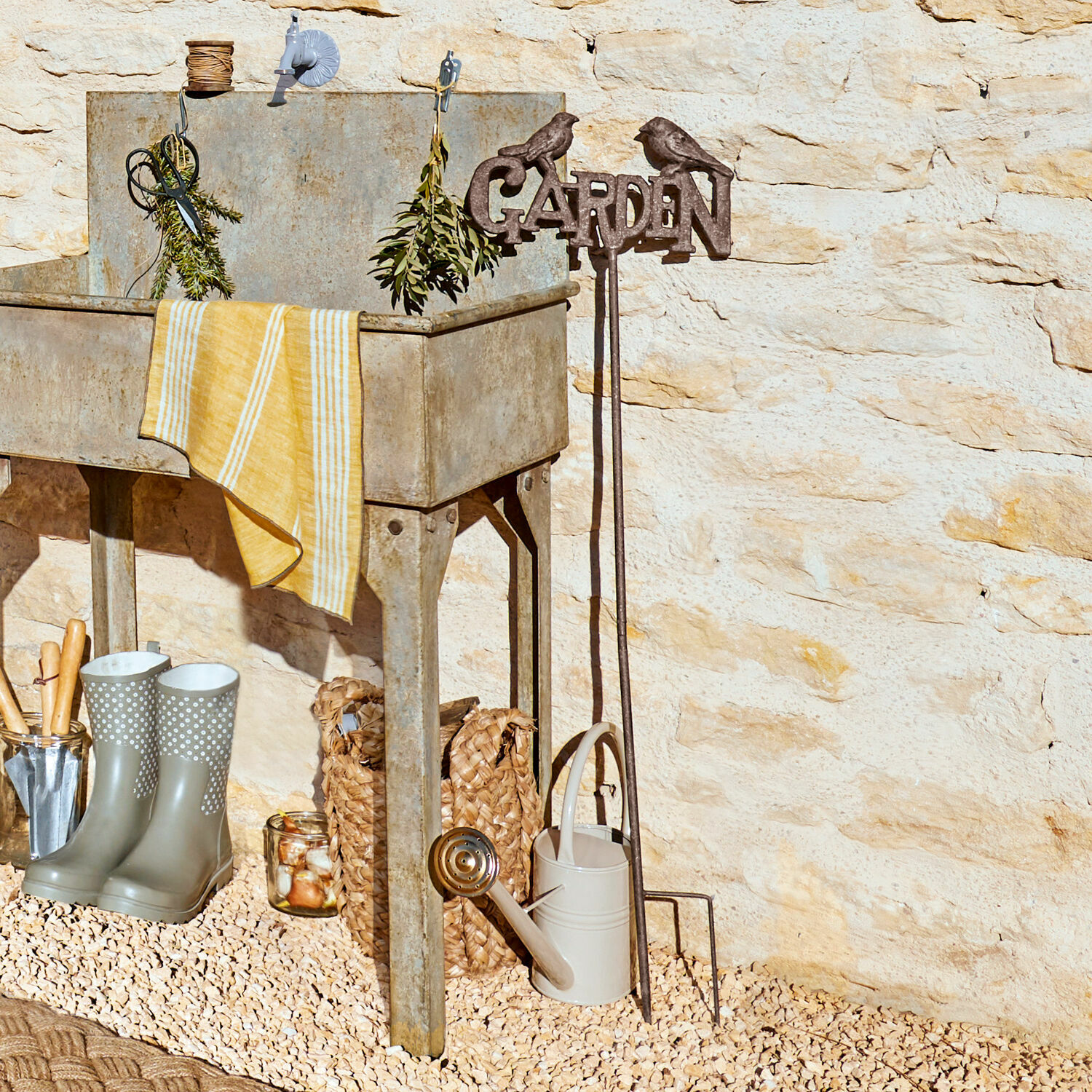 Gartenstab Girra Metall-Gartenspüle mit gelbem Tuch und Gartengeräten vor Steinmauer im Aussenbereich