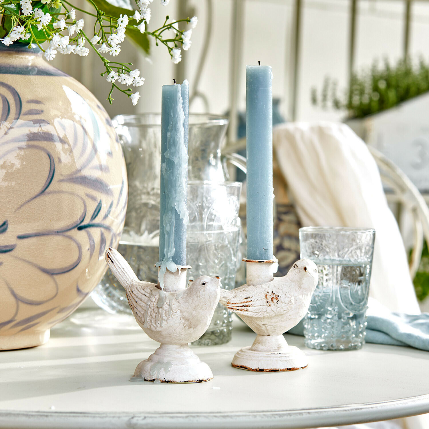 Weiße Vogel-Kerzenhalter mit blauen Kerzen auf hellem Tisch mit Blumendekor