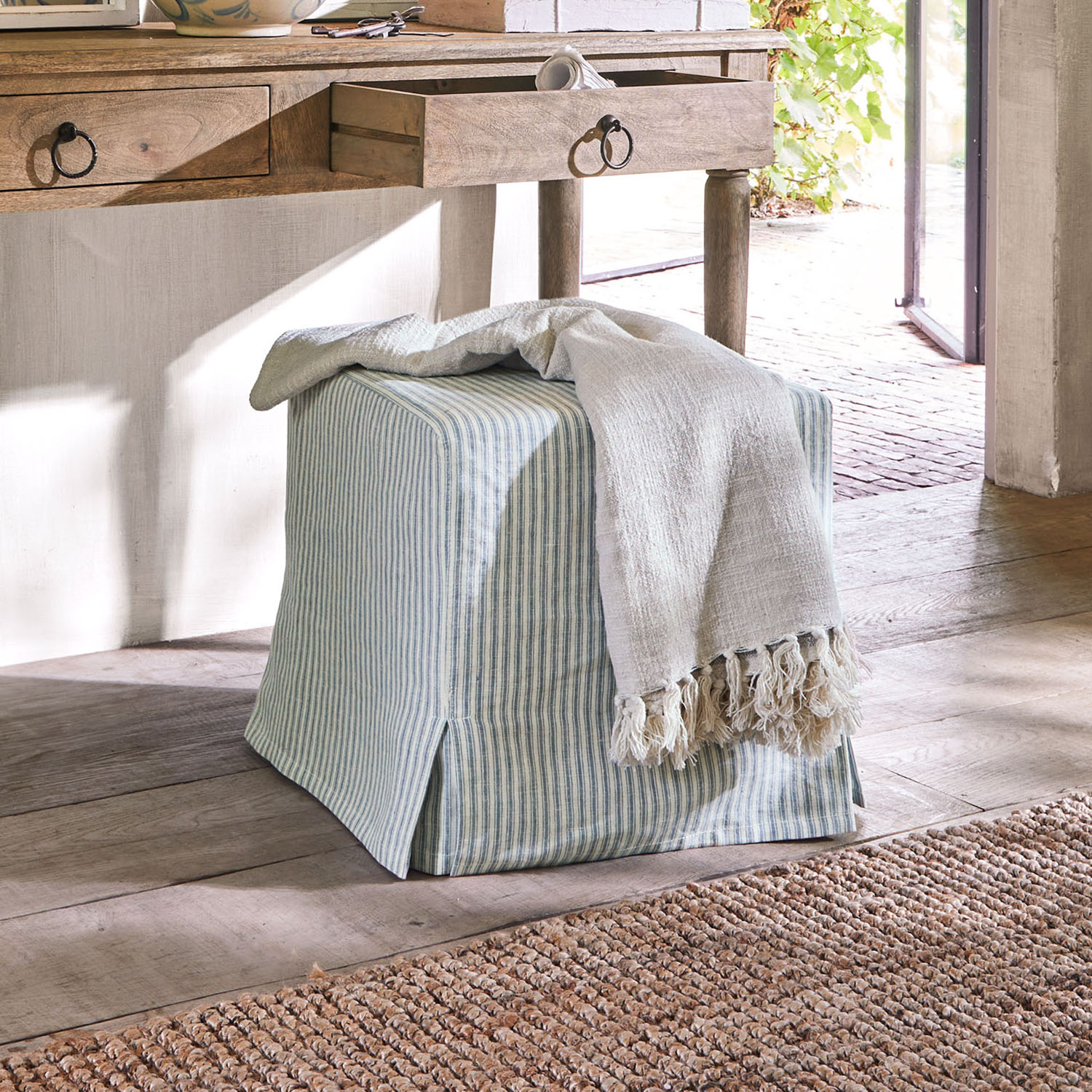 Quadratischer Baumwoll-Pouf mit blau-weiß gestreiftem Bezug und hellem Fransenplaid auf Holzboden