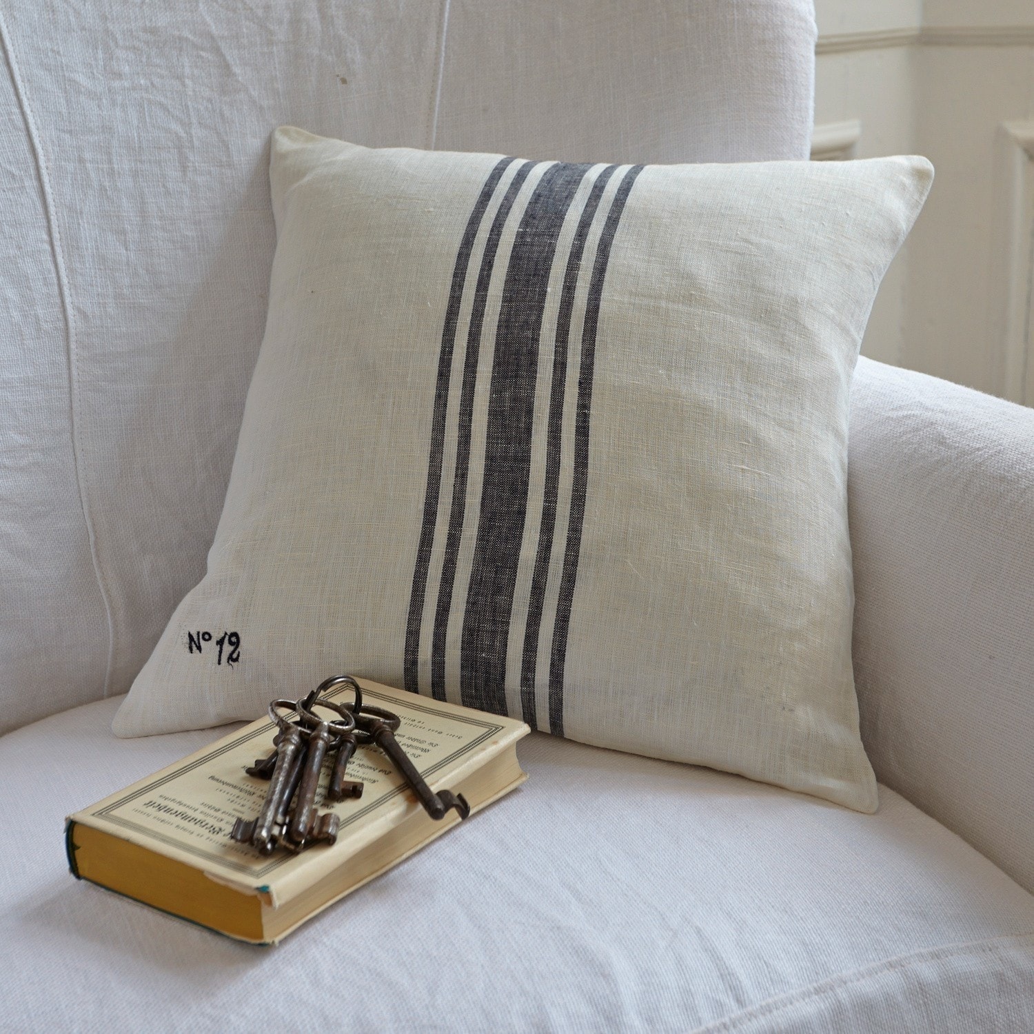 Kissen Lucian Dekoratives Kissen aus Leinen in Creme mit schwarzen Streifen auf weissem Sofa mit Buch und Schlüsseln