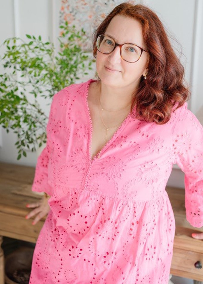 Frau in rosa Kleid mit Lochstickerei und zartem Muster, lehnt an einem Holztisch, im Hintergrund eine Pflanze.