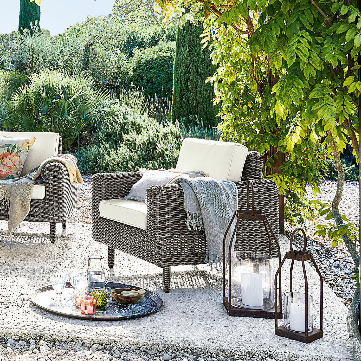 Sessel Bardou Gartenfauteuil aus grauem Rattan mit cremefarbenen Kissen und dekorativen Laternen auf der Terrasse