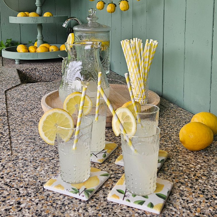 Gläser mit Limonade und Zitronenscheiben auf einer Steinplatte mit Papierstrohhalmen sowie einem Tablett mit Zitronen