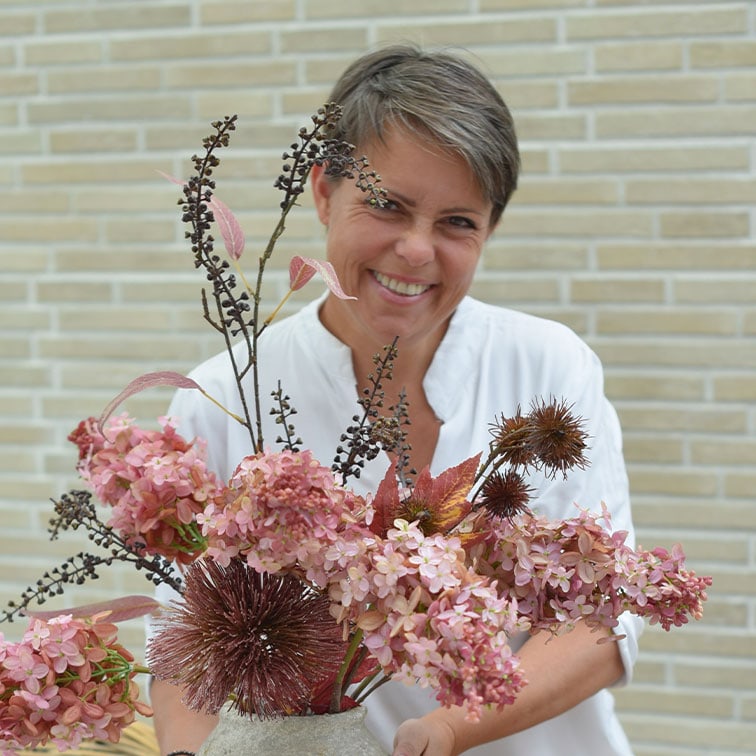 Frau in weissem Hemd hält Vase mit rosa und braunen getrockneten Dekoblumen