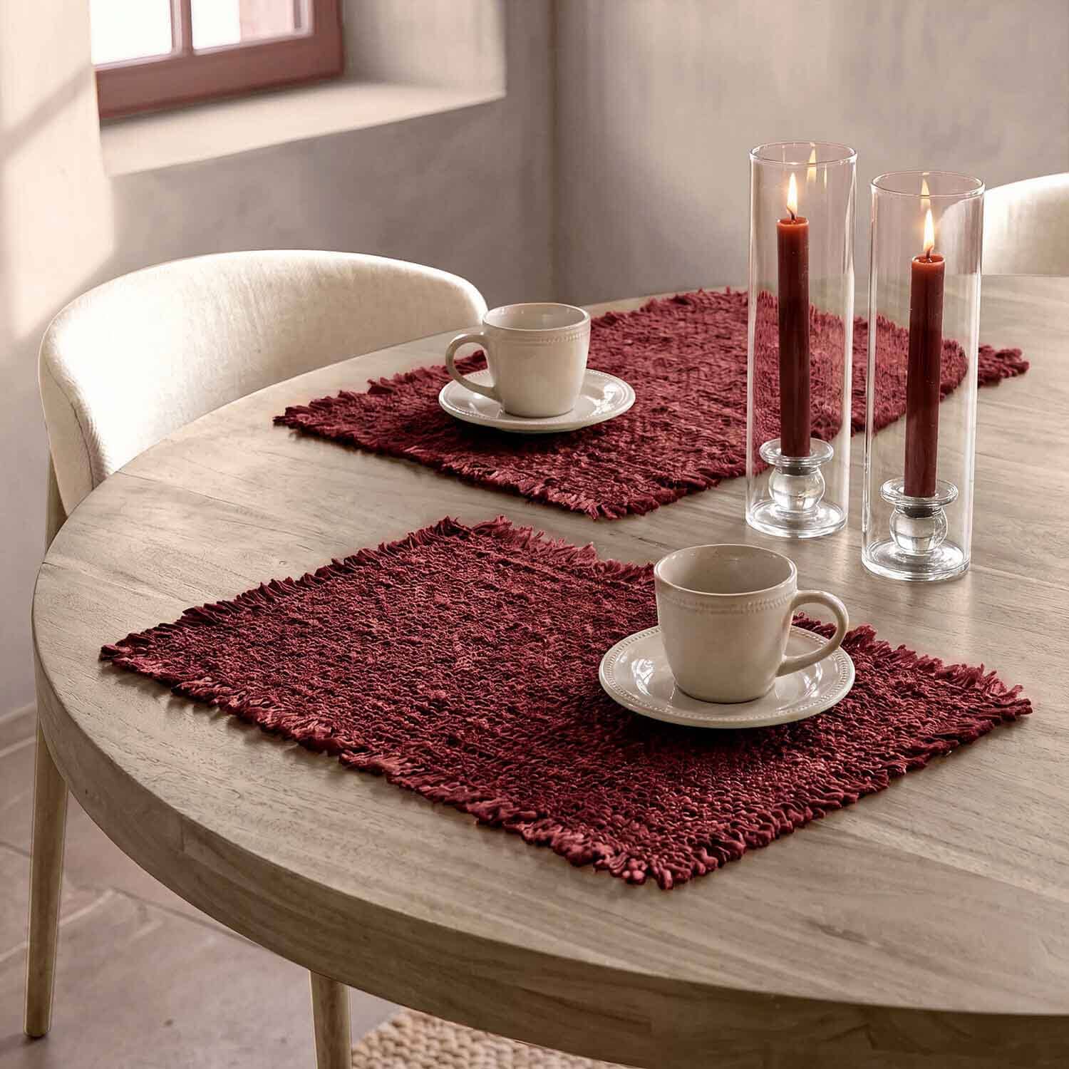 Holztisch mit bordeauxfarbenen Platzsets, weissen Tassen und roten Kerzen in Glaswindlichtern