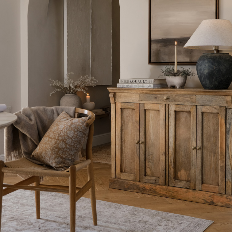 Holzkommode und Stuhl mit beigem Kissen im Wohnzimmer mit Lampe und Kerze, skandinavischer Stil