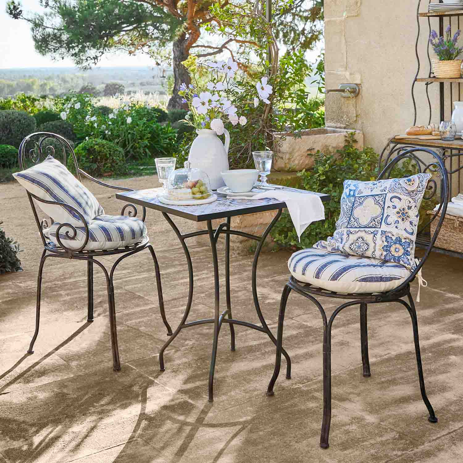 Metalltisch mit quadratischer Platte und zwei Stühle mit blauen Kissen auf sonniger Gartenterrasse