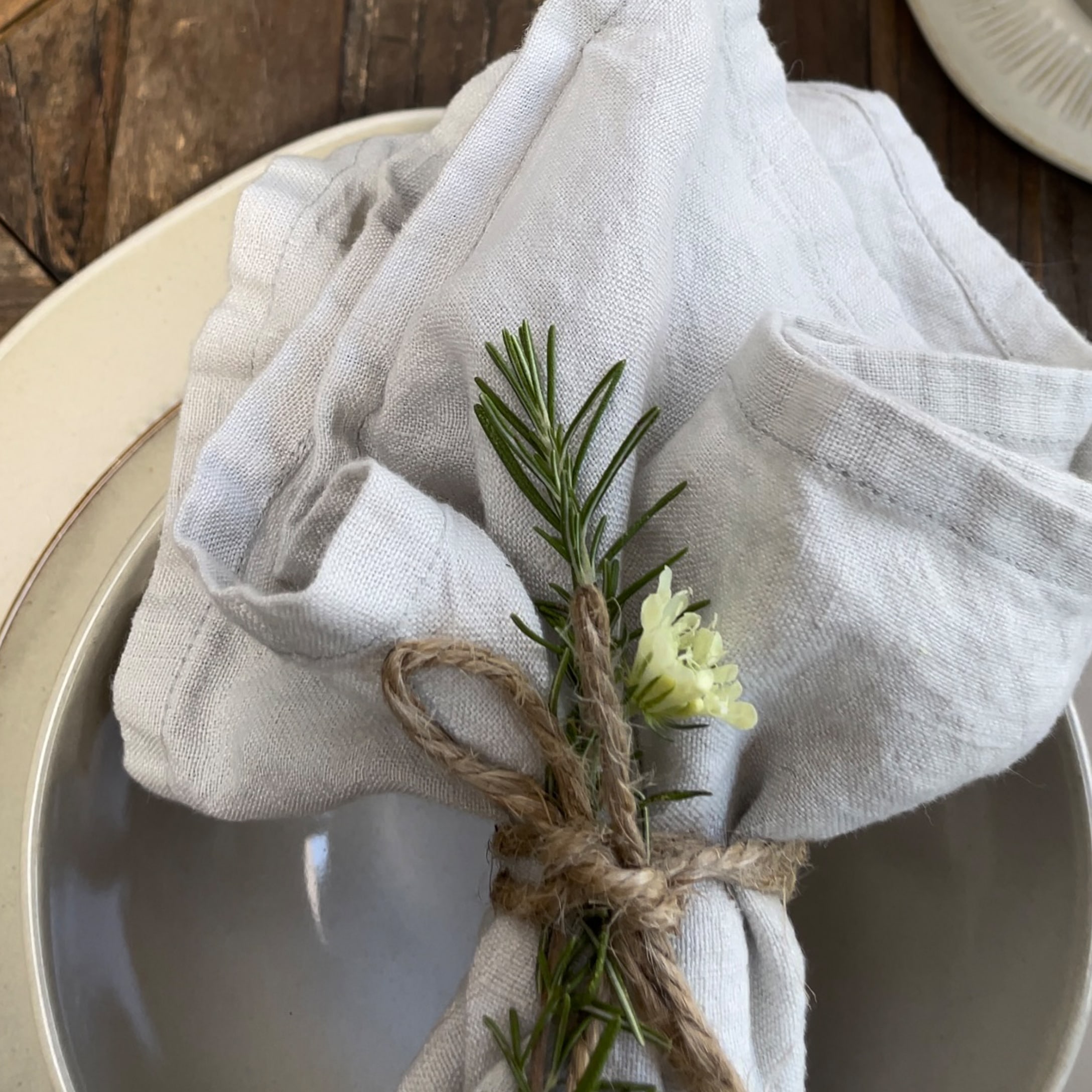 Weisse Leinenserviette mit Rosmarinzweig und Blüte auf Keramikteller dekorativ gebunden