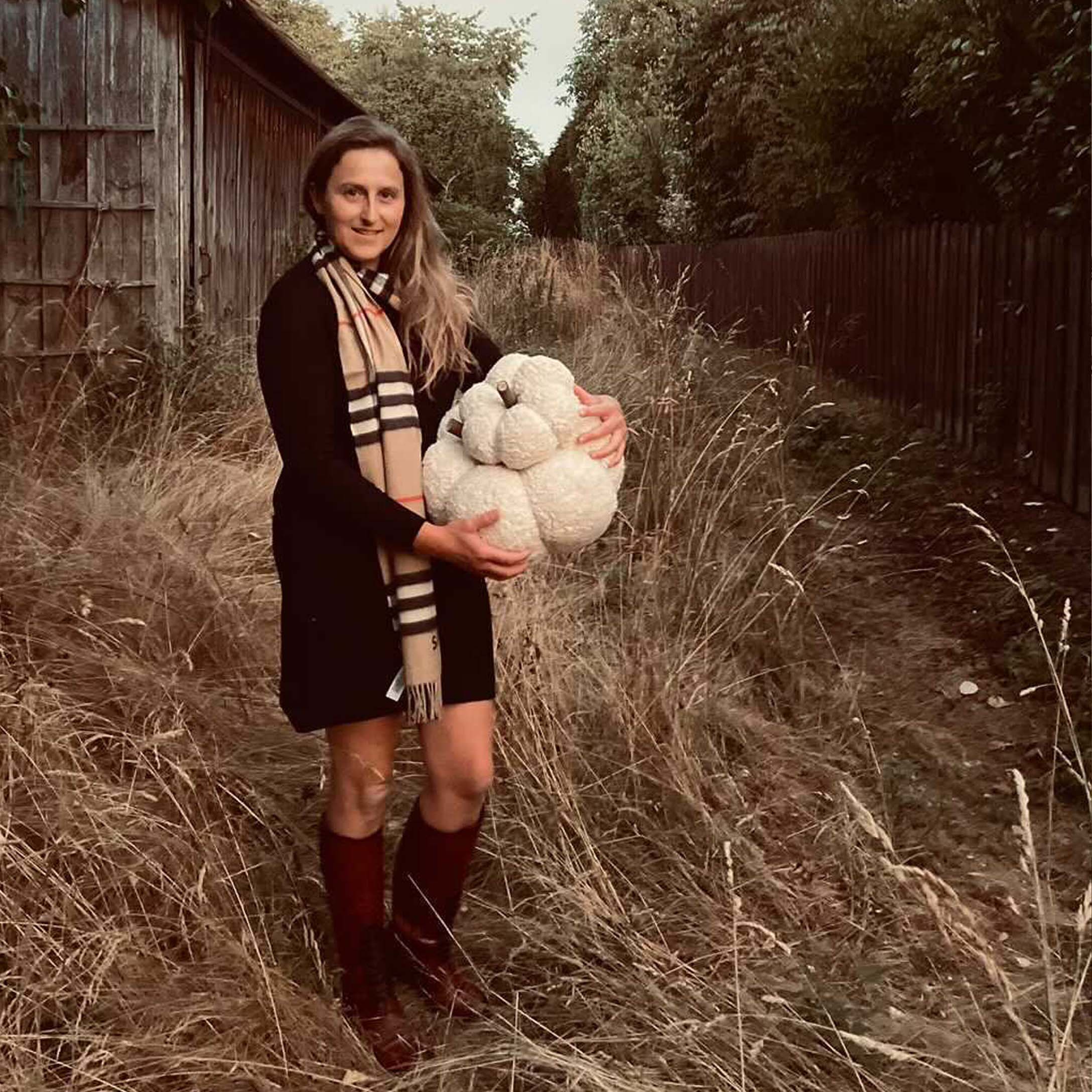 Frau im schwarzen Kleid und Schal hält einen weissen Dekokürbis auf einer herbstlichen Wiese.