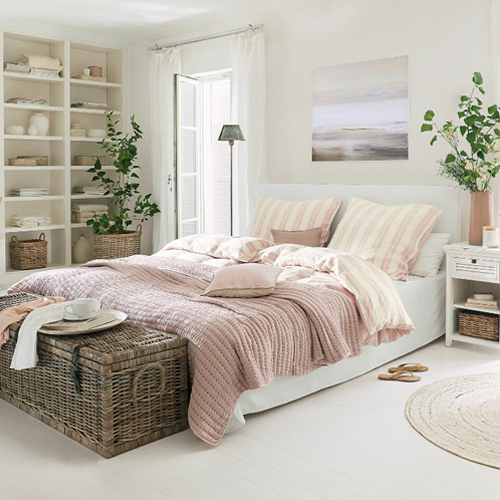 Gemütliches Schlafzimmer mit weisser Bettwäsche und rosa Decke, natürlichen Dekorationen und Korbtruhe