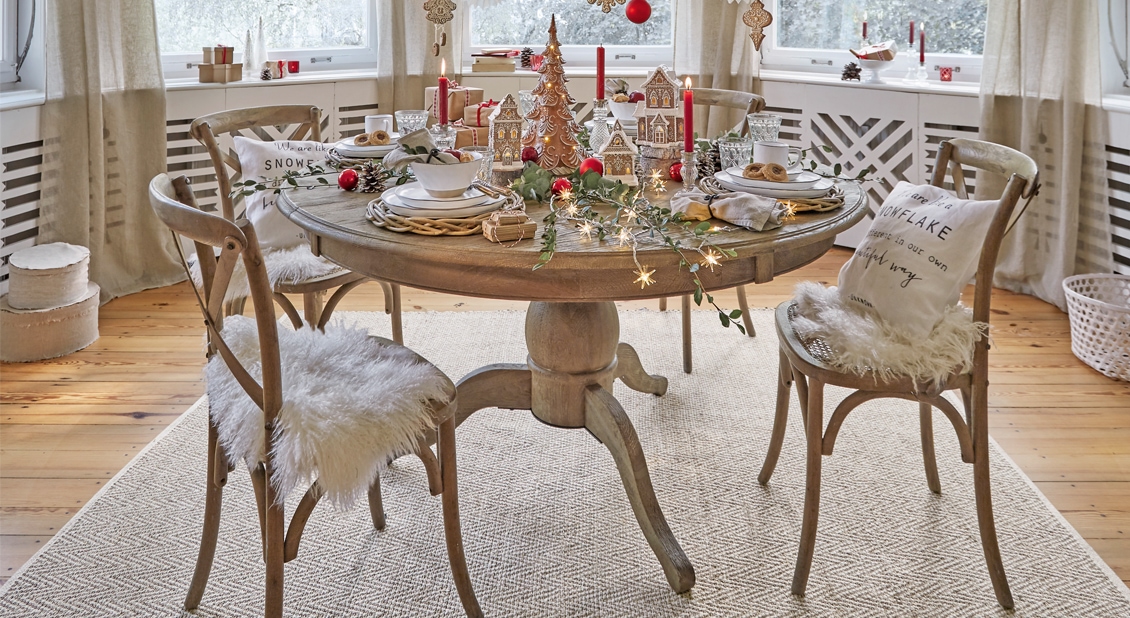 Runder Holztisch mit Weihnachtsdekoration und hellen Stühlen mit Fellkissen in einem gemütlichen Raum