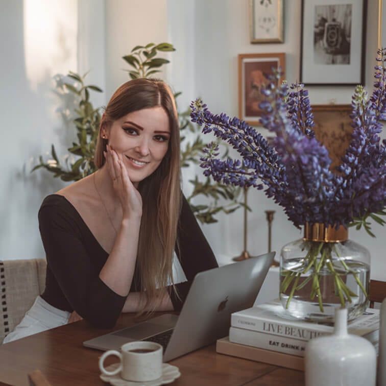 Frau arbeitet am Laptop an Holztisch mit lila Blumenstrauss und Kaffeetasse